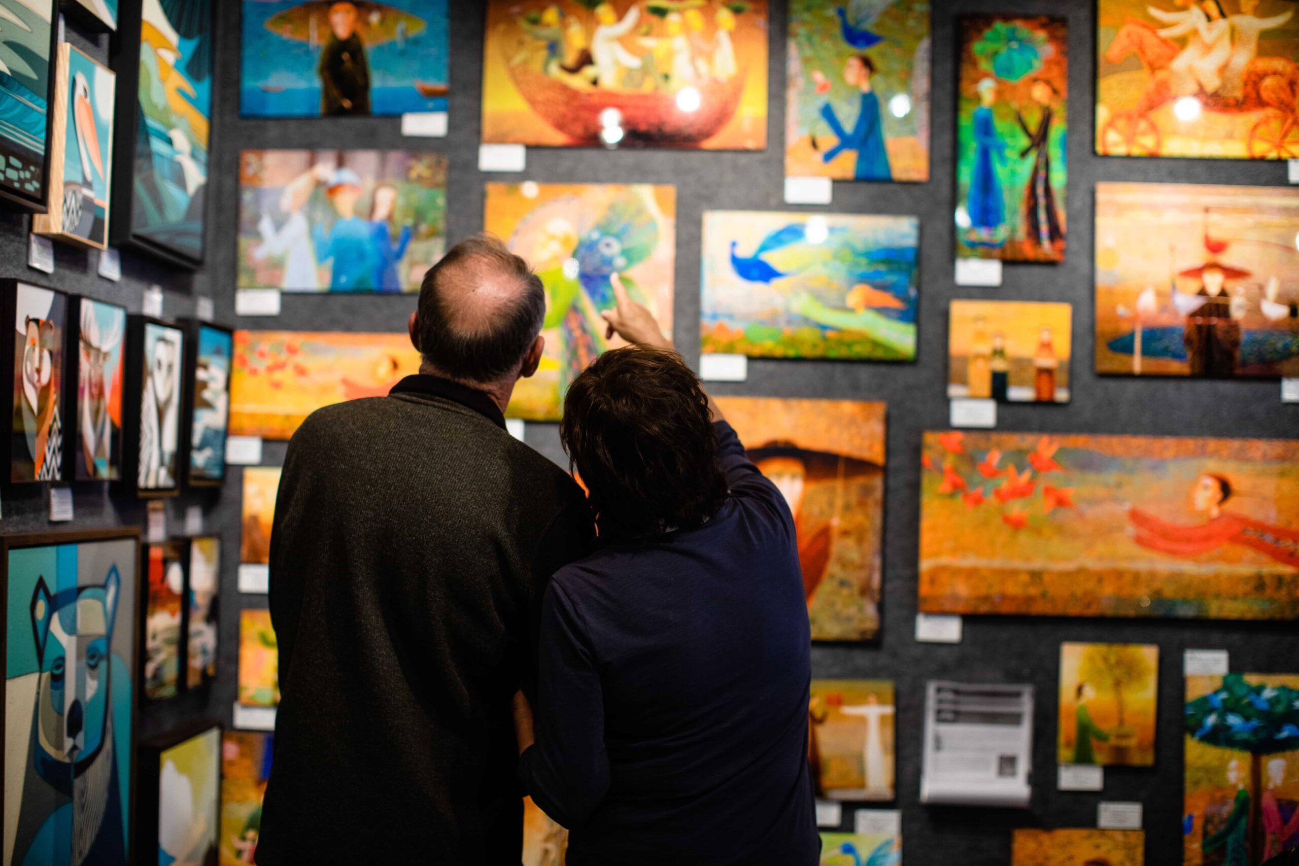 A couple is looking up at a wall full of colorful paintings hung in a gallery.