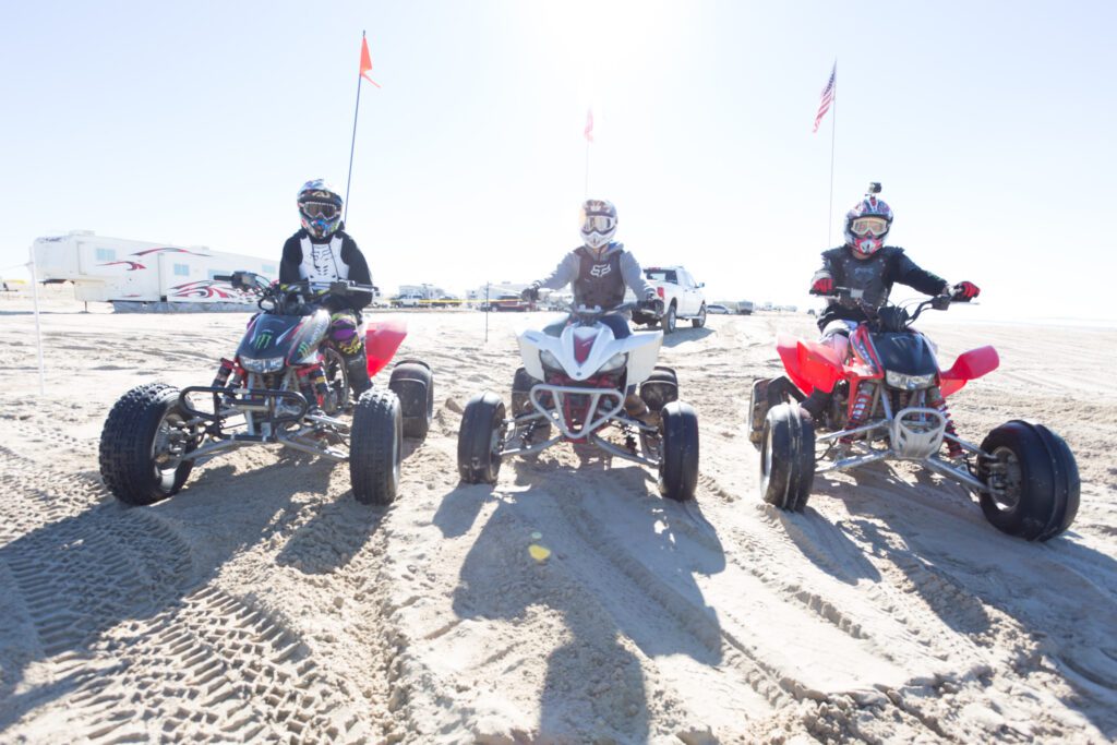 3 young adults on ATV's driving alongside one another on beach dunes.