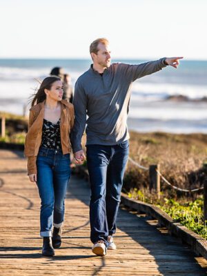 A man and woman walking on a wooden path by the ocean.