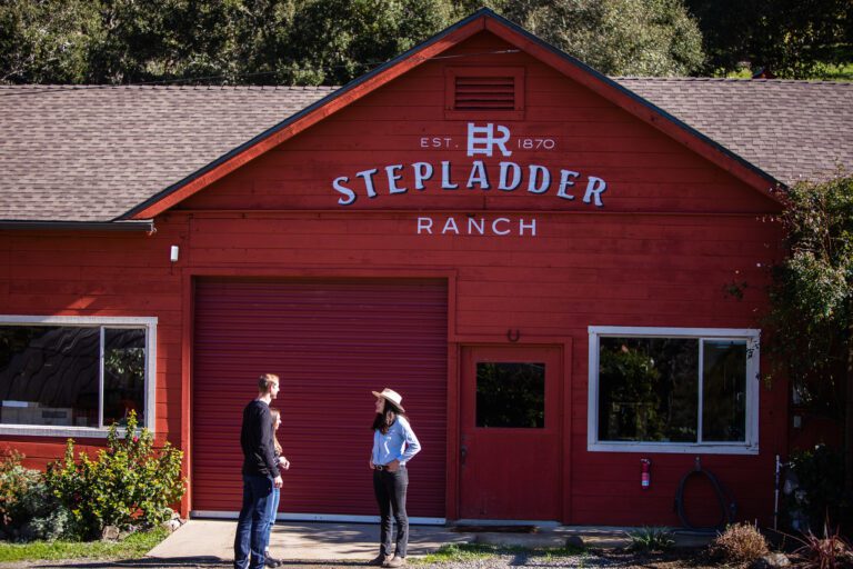 The image features a few people standing outside a building with a sign that reads "EST 1870 STEP LADDER RANCH."
